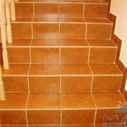 Schody wyłożone brązowymi, kwadratowymi płytkami ceramicznymi z jasną fugą, widok z góry, drewniana balustrada po lewej stronie.